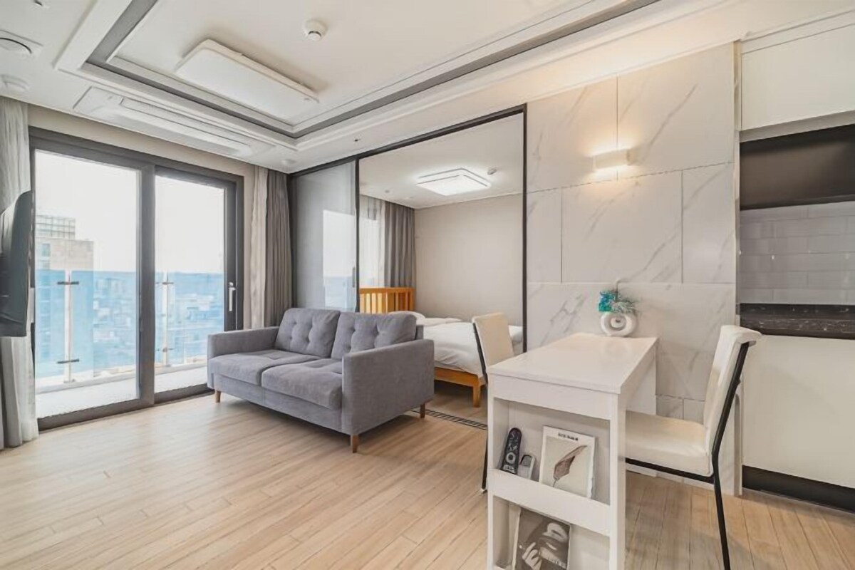 A modern hotel room with a large bed, a grey sofa, a dining table, and a view of the city.