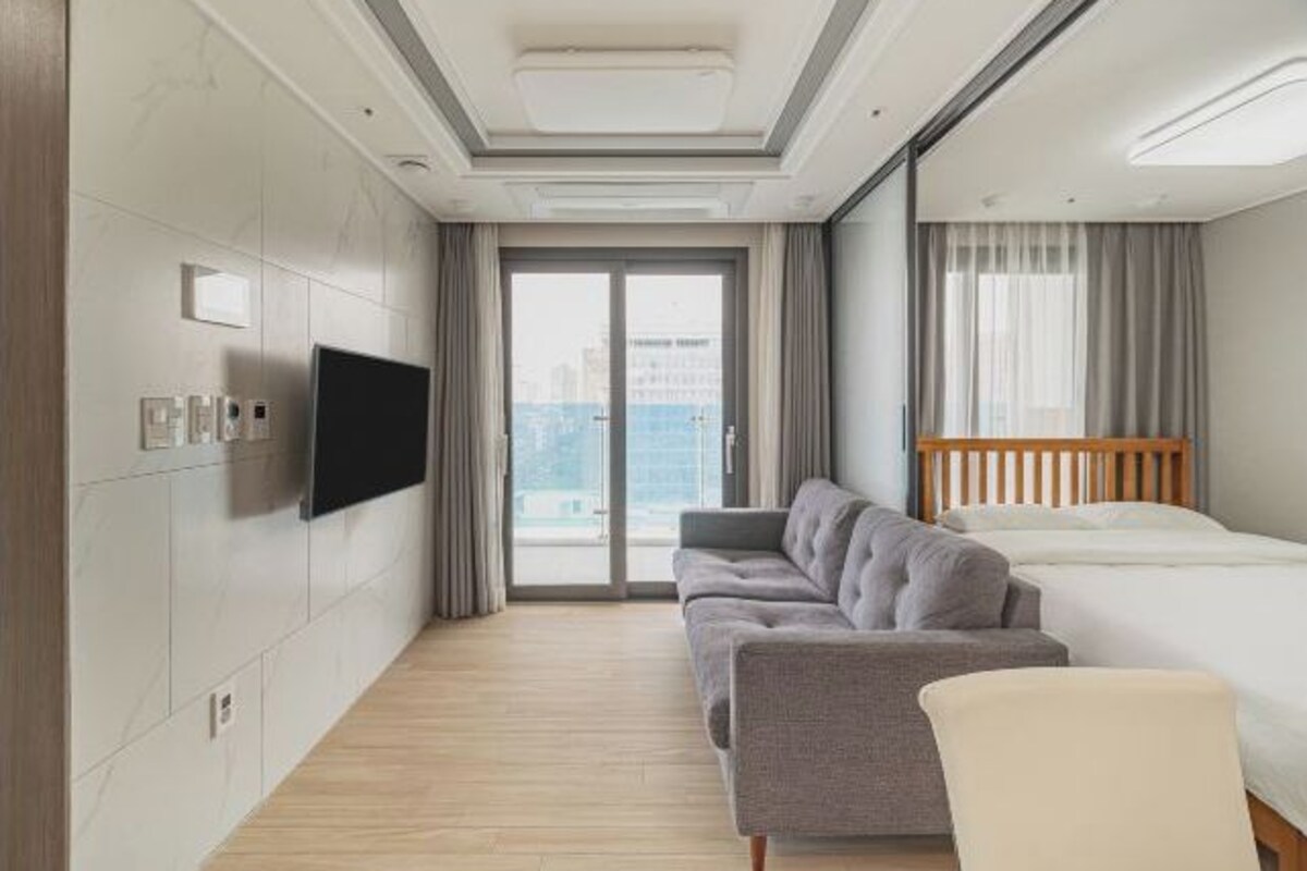 A modern hotel room with a sofa, bed, TV, and large windows.
