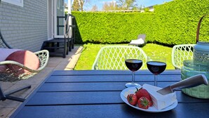 Een terras met een tafel gedekt voor twee personen, compleet met een bord aardbeien, een kaasmesser, twee glazen rode wijn en een groene haag op de achtergrond.