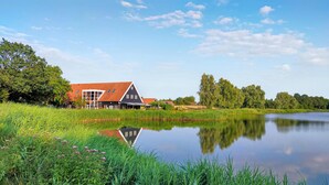Een groot gebouw met een rood dak en meerdere ramen, gelegen naast een rustig meer met weelderige begroeiing en bomen op de achtergrond.