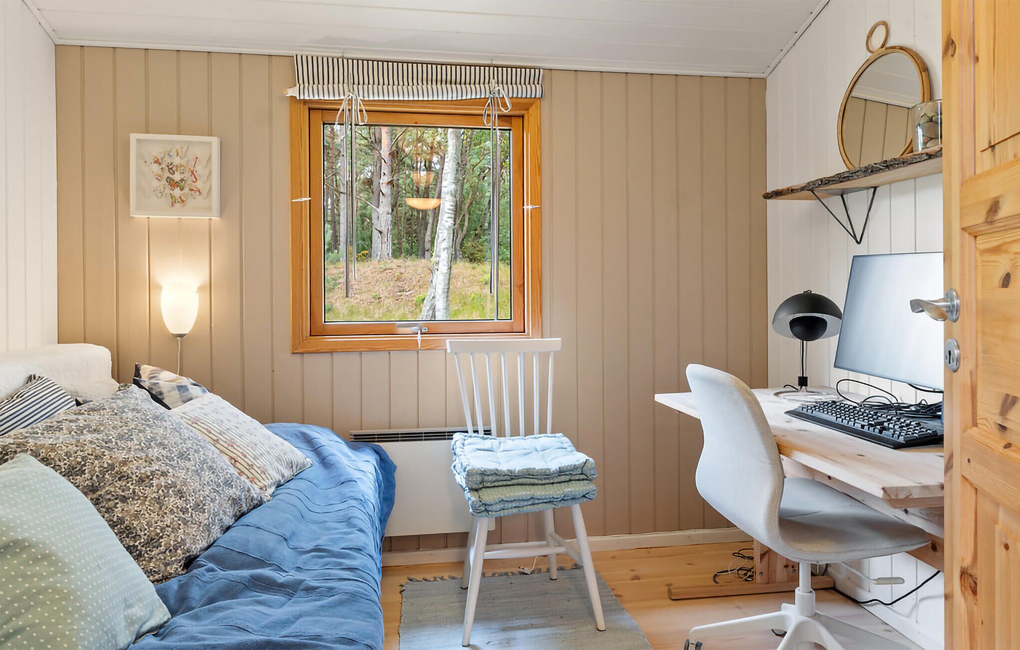 Een slaapkamer met een bed, een bureau met een computer, een stoel, een raam met uitzicht op bomen en een wandlamp.