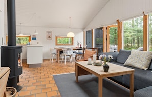 A cozy cabin interior with a central fireplace, a dining area, and a seating area with a view of the forest. - 3 bedroom lovely home in Nexø (Nexø)