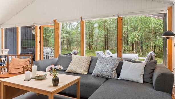 Ein gemütliches, modernes Wohnzimmer mit einem großen, grauen Ecksofa, einem Holztisch und Blick auf einen Wald durch große Fenster.