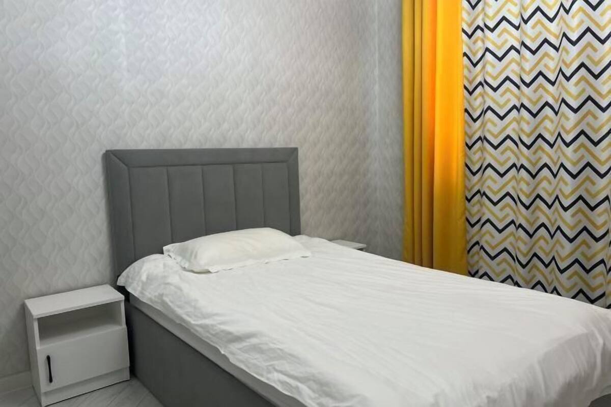 A single bed with a grey headboard, white bedding, a small white nightstand with a drawer, and a patterned curtain.