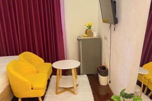 A hotel room with a bed, a yellow armchair, a small white table, a television mounted on the wall, a small cabinet with a plant, and a red curtain. - Durian Family Hotel (Bangkok)