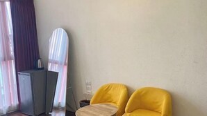 Ein Zimmer mit einem Bett, einem kleinen Tisch, zwei gelben Stühlen, einem Spiegel und einem Fenster mit Vorhängen.