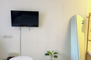 A room with a mounted television, a surfboard leaning against the wall, and a small plant. - Durian Family Hotel (Bangkok)