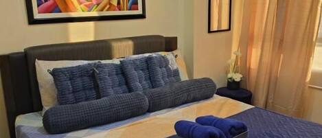 A neatly made bed with a blue quilt, pillows, and a blue blanket. There is a colorful abstract painting on the wall above the bed.