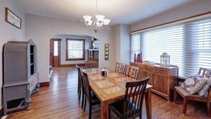 A spacious dining area with a wooden table and chairs, a chandelier, and a large window with blinds. - The Charming Chestnut (Arkansas City)