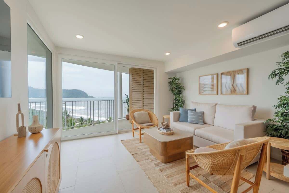 Uma sala de estar moderna com sofá, mesa de centro e cadeiras de vime. Possui uma varanda com vista para o mar.