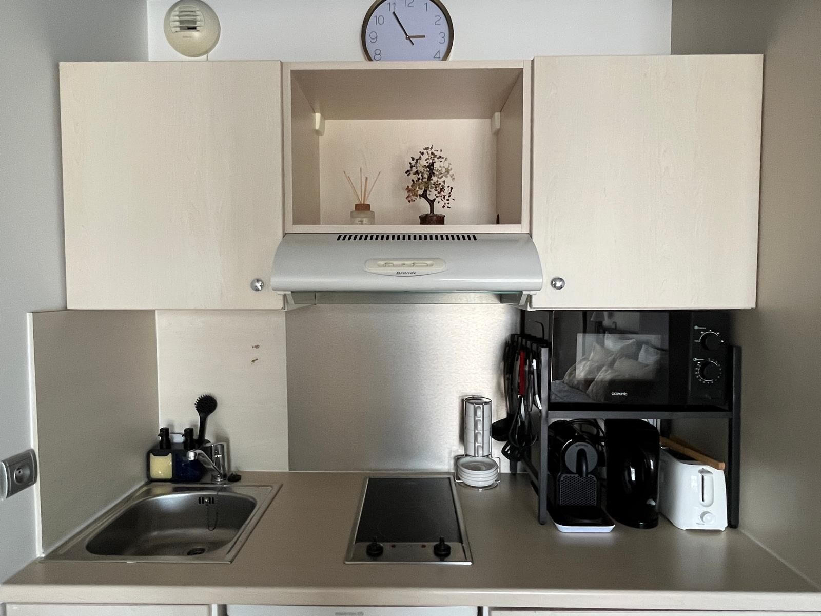 A compact kitchen with a sink, stove, toaster, and a clock on the wall.