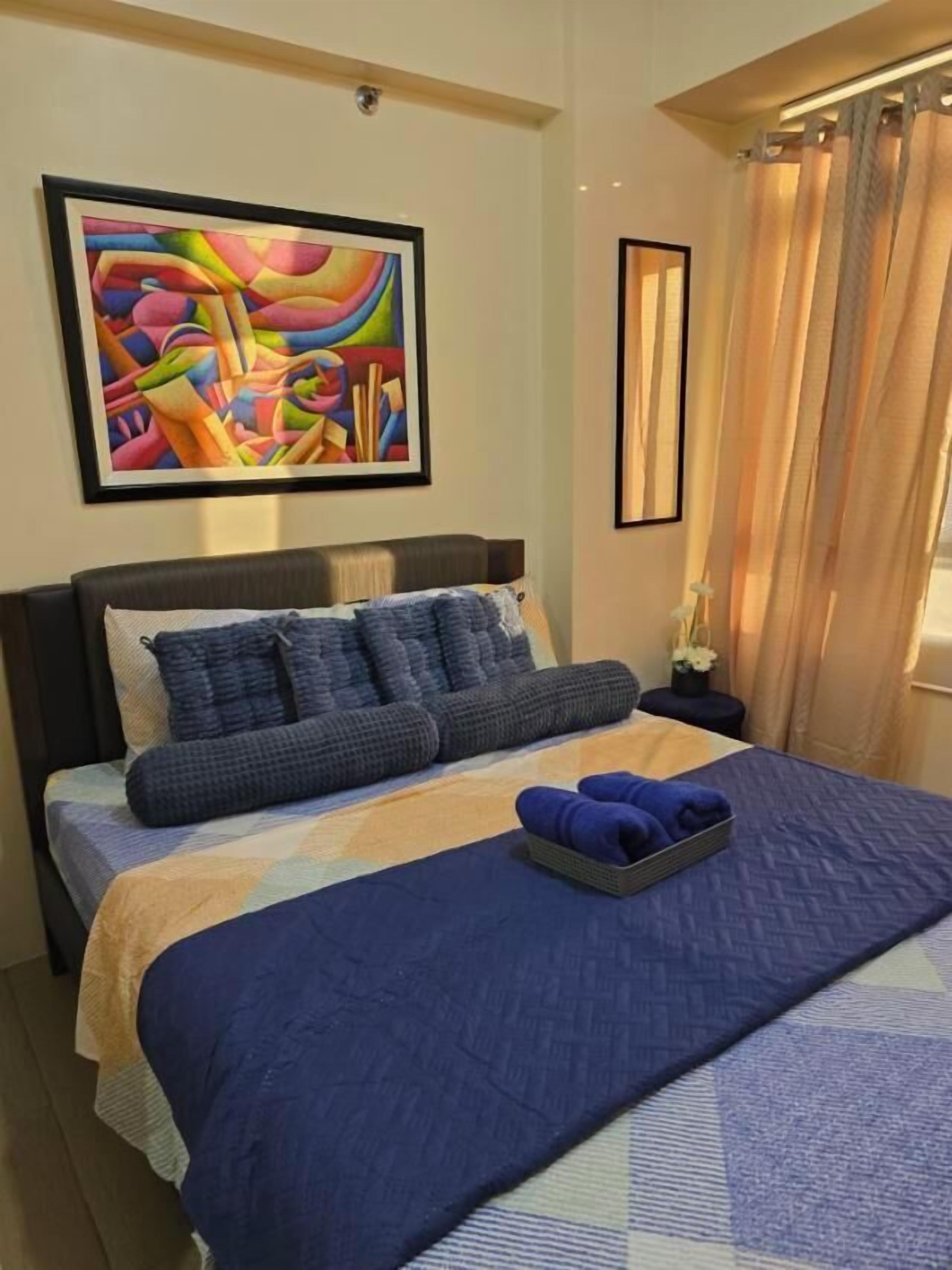 Un llit ben fet amb un llençol blau, almoinades i una manta blava. Hi ha un quadre abstracte i colorit a la paret sobre el llit.