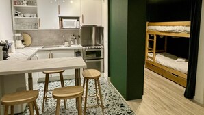 Een compacte keuken met een magnetron, fornuis en wastafel, een eethoek met krukken, en een stapelbed met een groene wand.