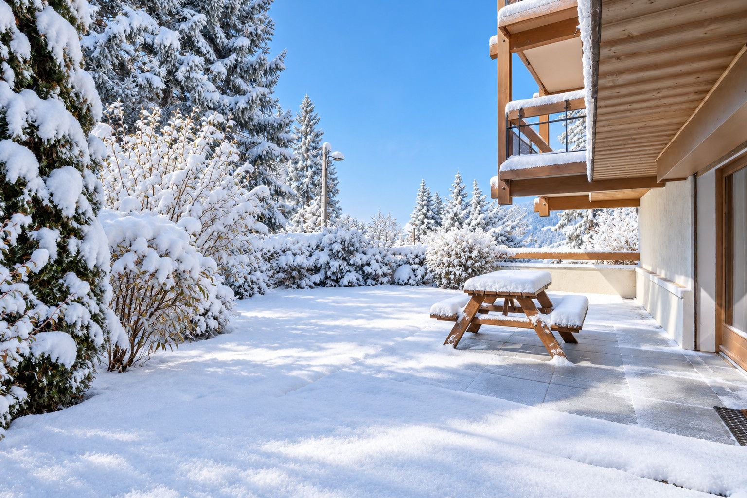 Un espace extérieur enneigé, avec un banc en bois et une table, entouré d’arbres chargés de neige.