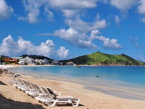 Une plage avec des chaises longues blanches alignées le long du rivage, un ciel bleu dégagé et une petite colline en arrière-plan.