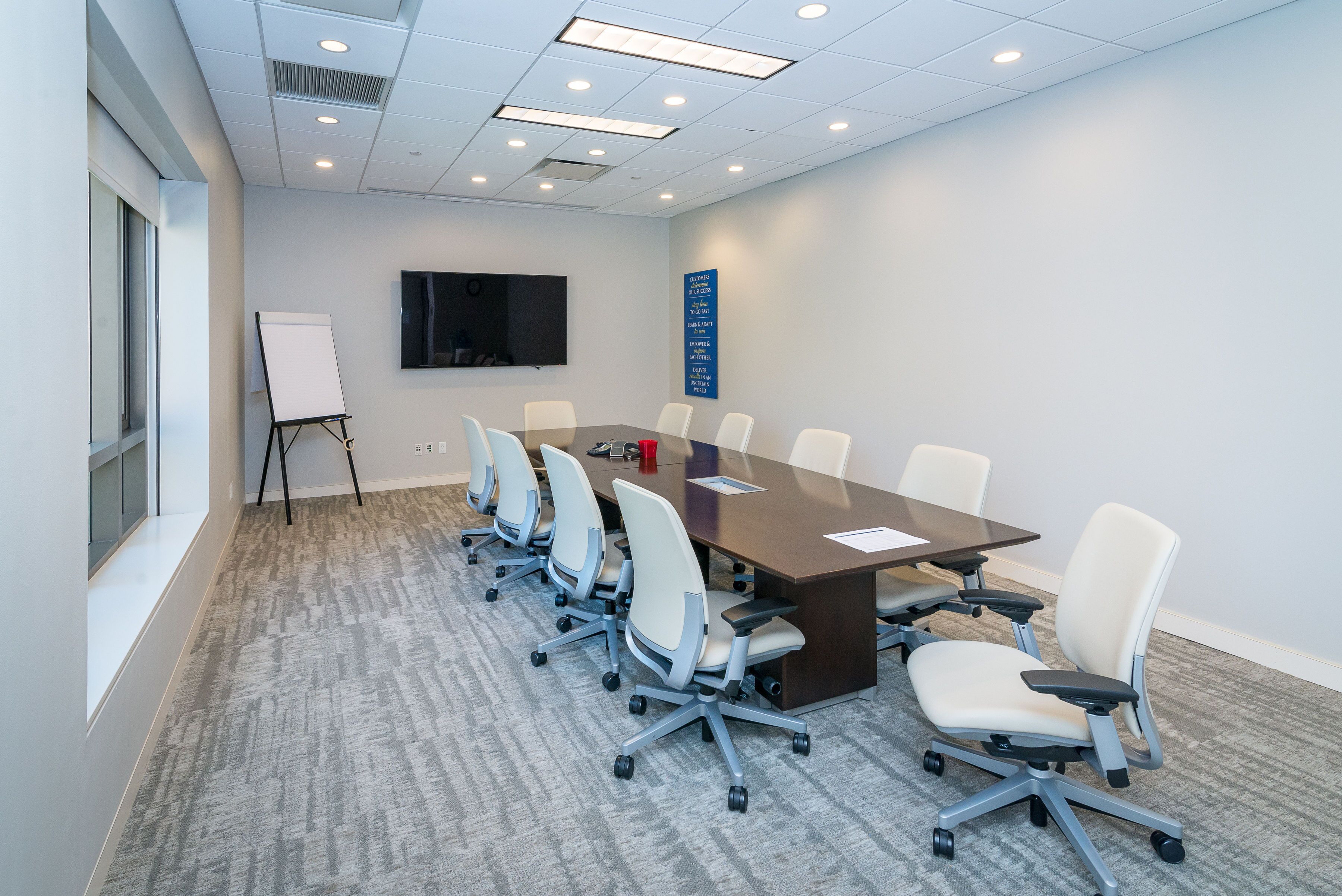 A conference room with a long table, six chairs, a whiteboard, and a TV on the wall.