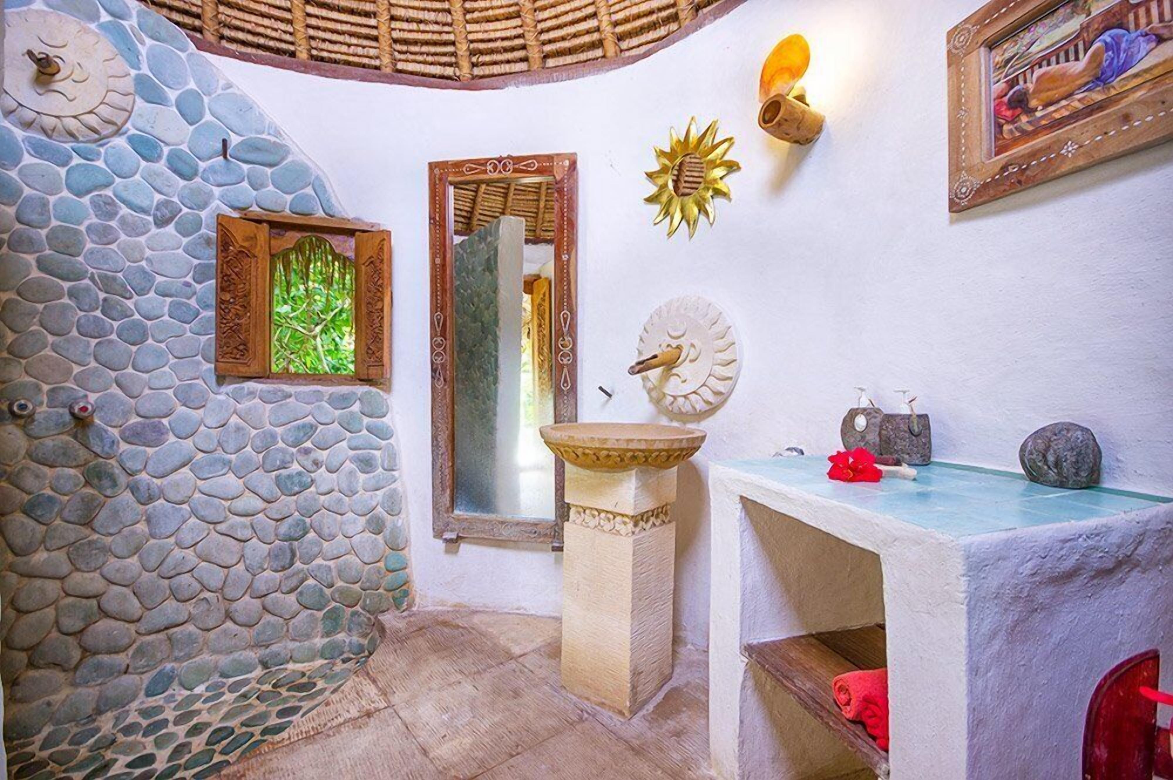 Casa de banho com parede de pedra, espelho com moldura de madeira, lavatório e candeeiro decorativo em forma de sol.