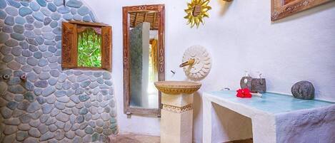 Casa de banho com parede de pedra, espelho com moldura de madeira, lavatório e candeeiro decorativo em forma de sol.