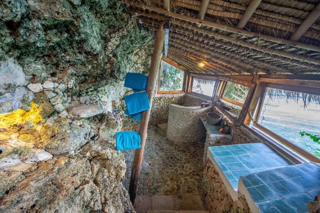 Una habitación de paredes de piedra con techo de paja, un banco de piedra y una ventana con vista a un cuerpo de agua.