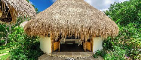 A thatched-roof hut with a stone-paved entrance, surrounded by lush greenery.