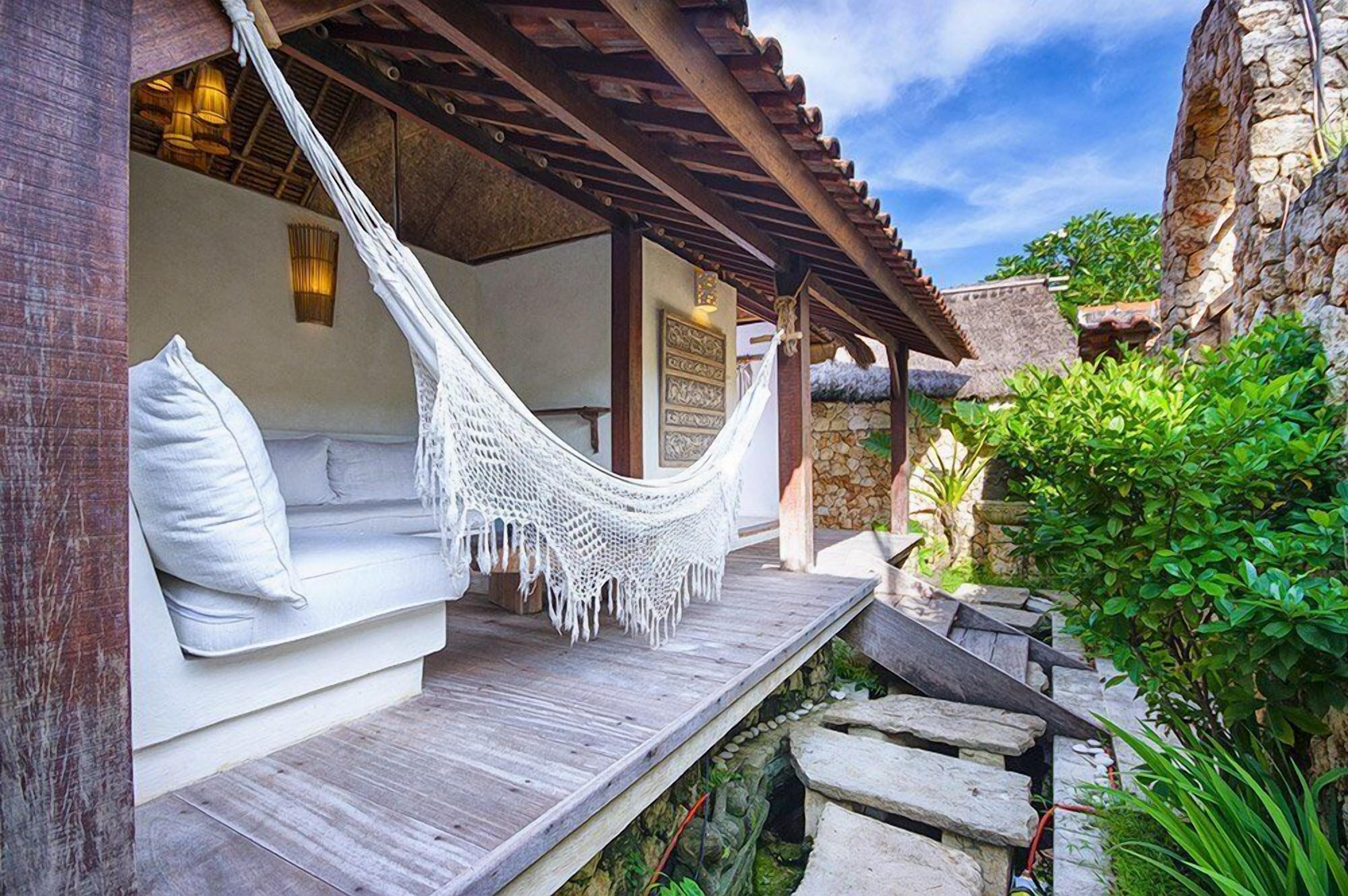 A hammock chair with a cushion, a wooden bench, and a stone wall.