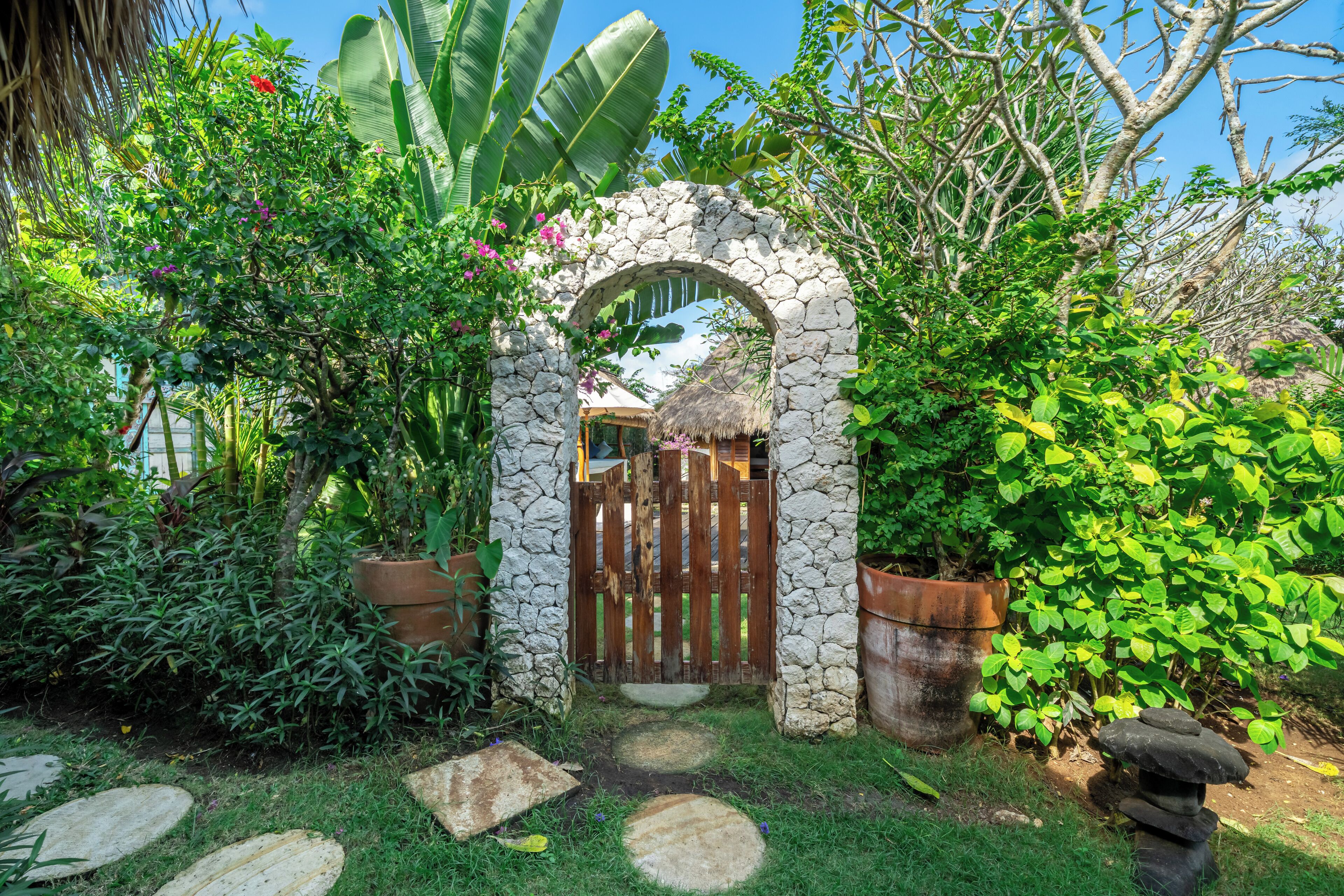 Un arc en pierre mène à une porte en bois, l’ensemble étant entouré d’une végétation luxuriante et de plantes en pot.