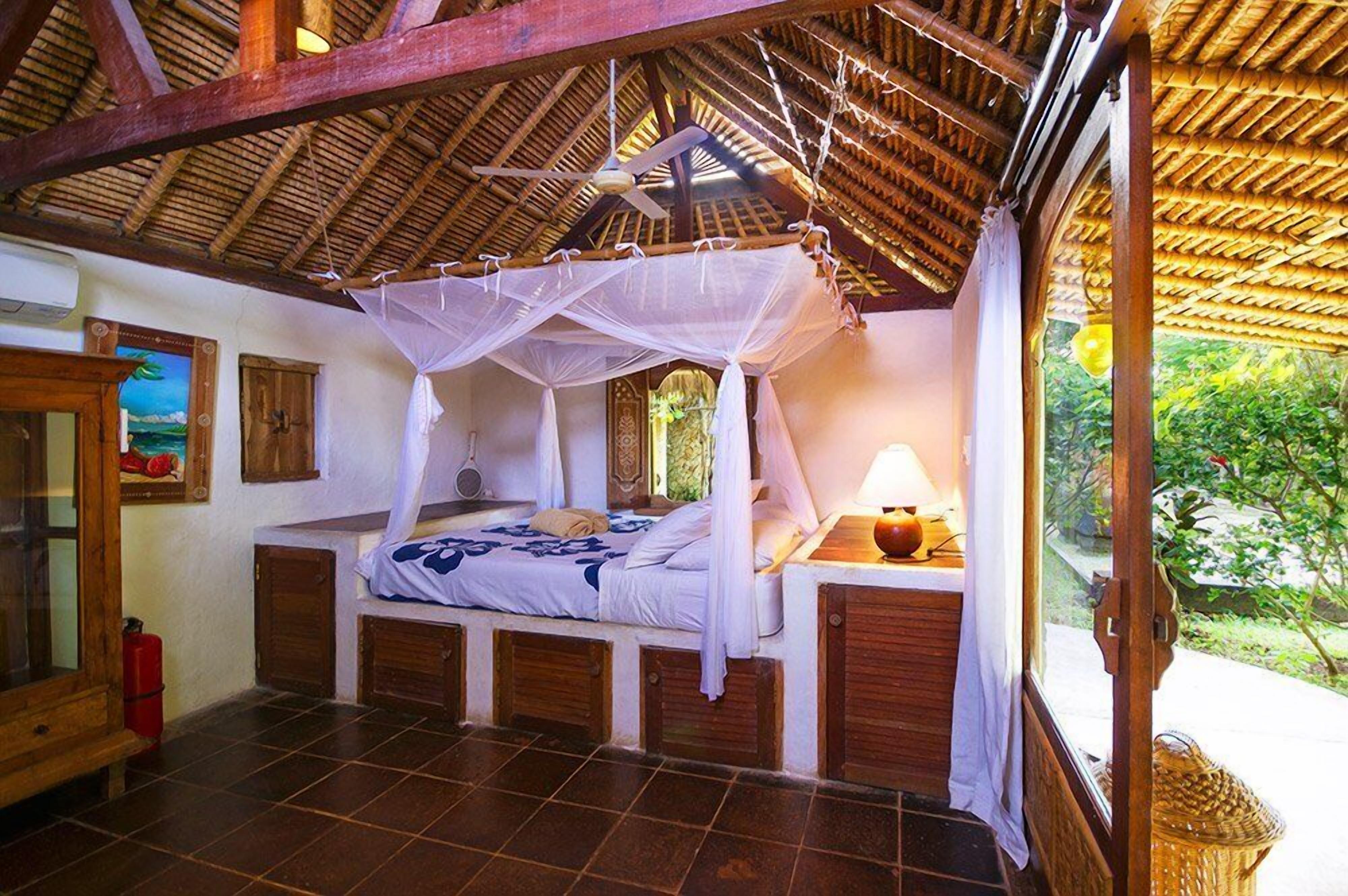 A bedroom with a bed under a canopy, wooden beams, a window with a view of greenery, and a bedside table with a lamp.
