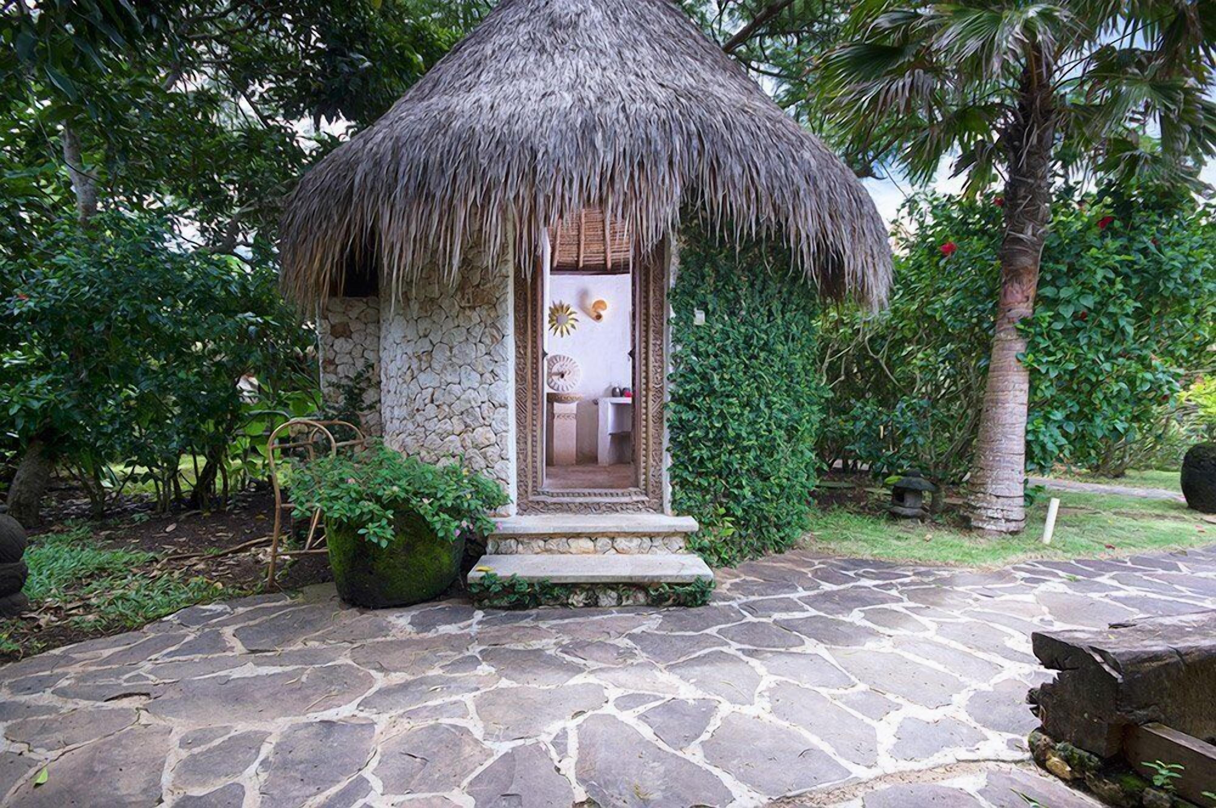 A thatched-roof structure with a stone base, surrounded by greenery and a stone-paved area.