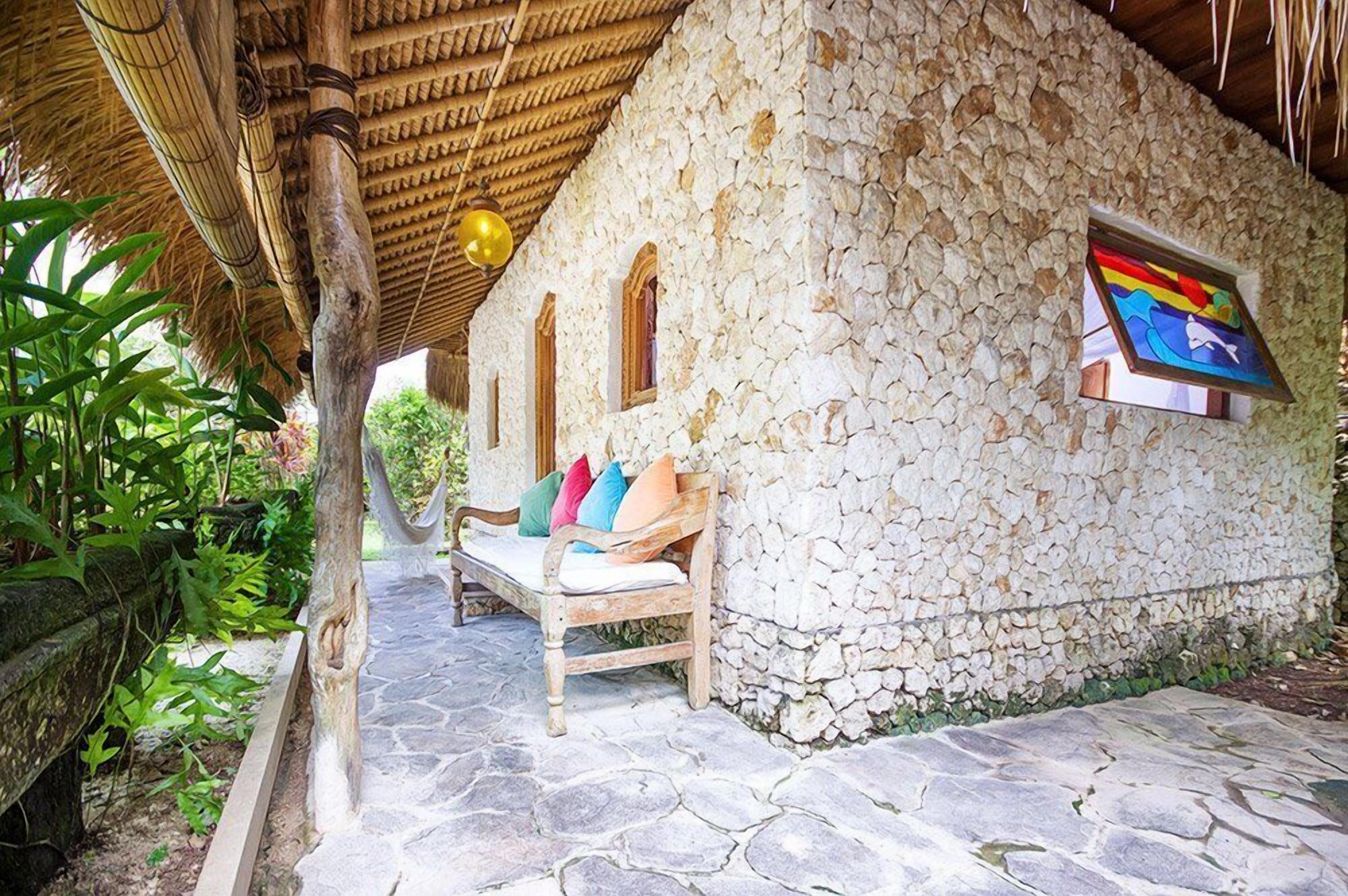 A stone-walled building with a thatched roof, a bench with cushions, and a framed picture on the wall.