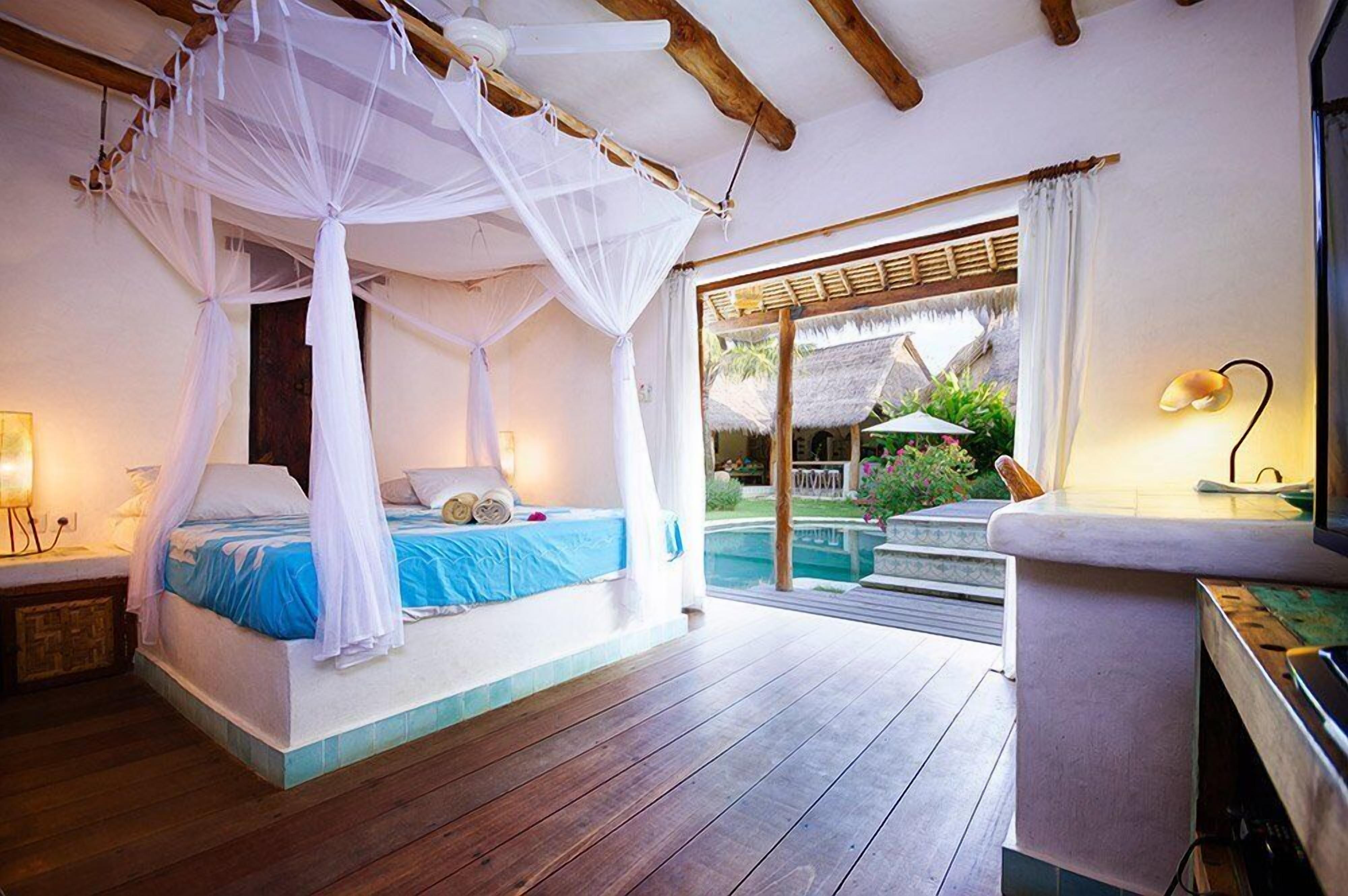 Ein Schlafzimmer mit einem Himmelsbett, ein Pool ist durch eine Glastür sichtbar, und ein kleiner Tisch mit einer Lampe.