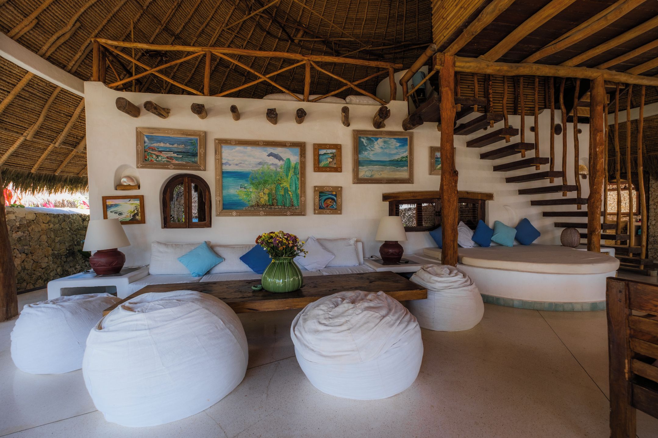 A room with a thatched roof, a sofa set, a dining table, and a staircase.