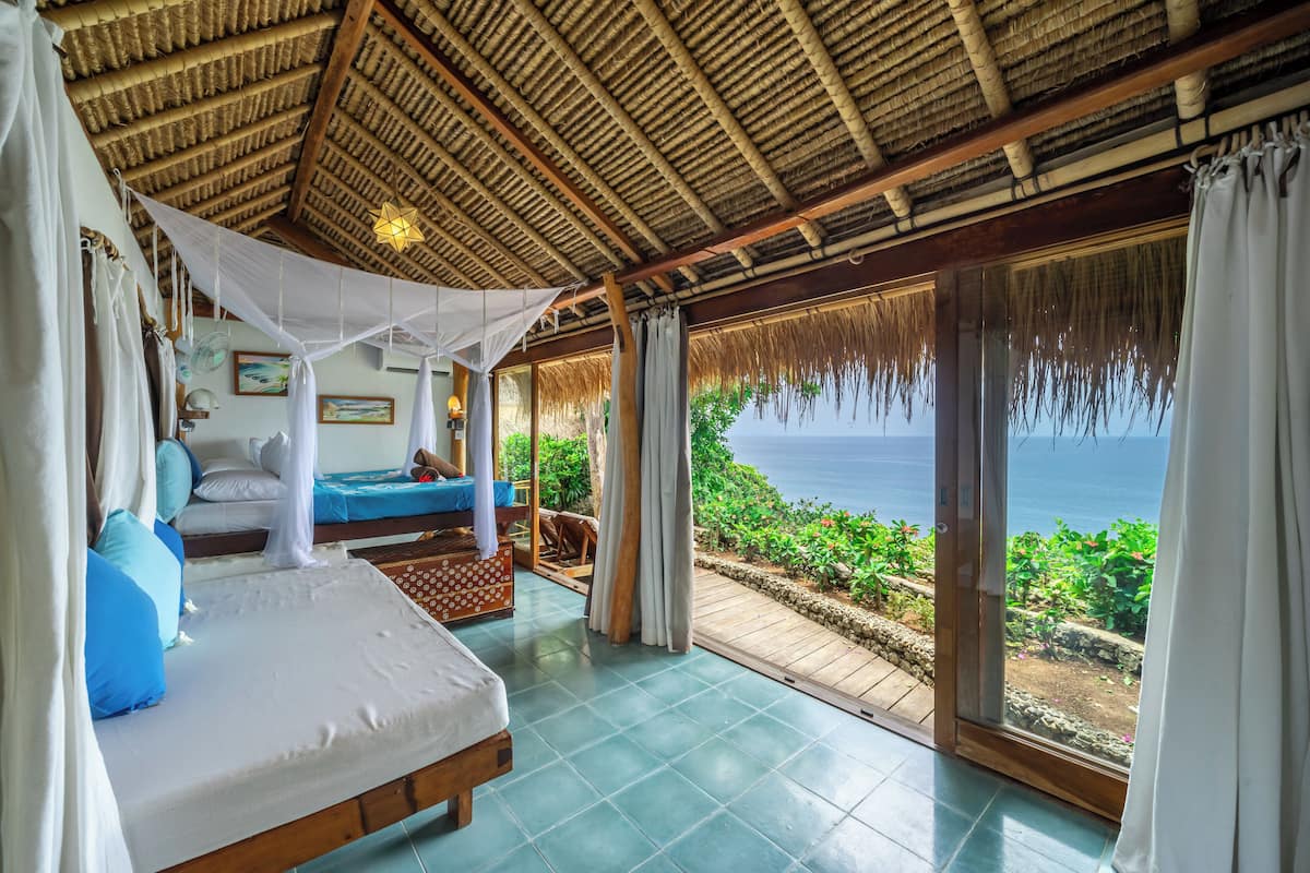 Een slaapkamer met een bed, nachtkastje, raam met uitzicht op zee en rieten dak.