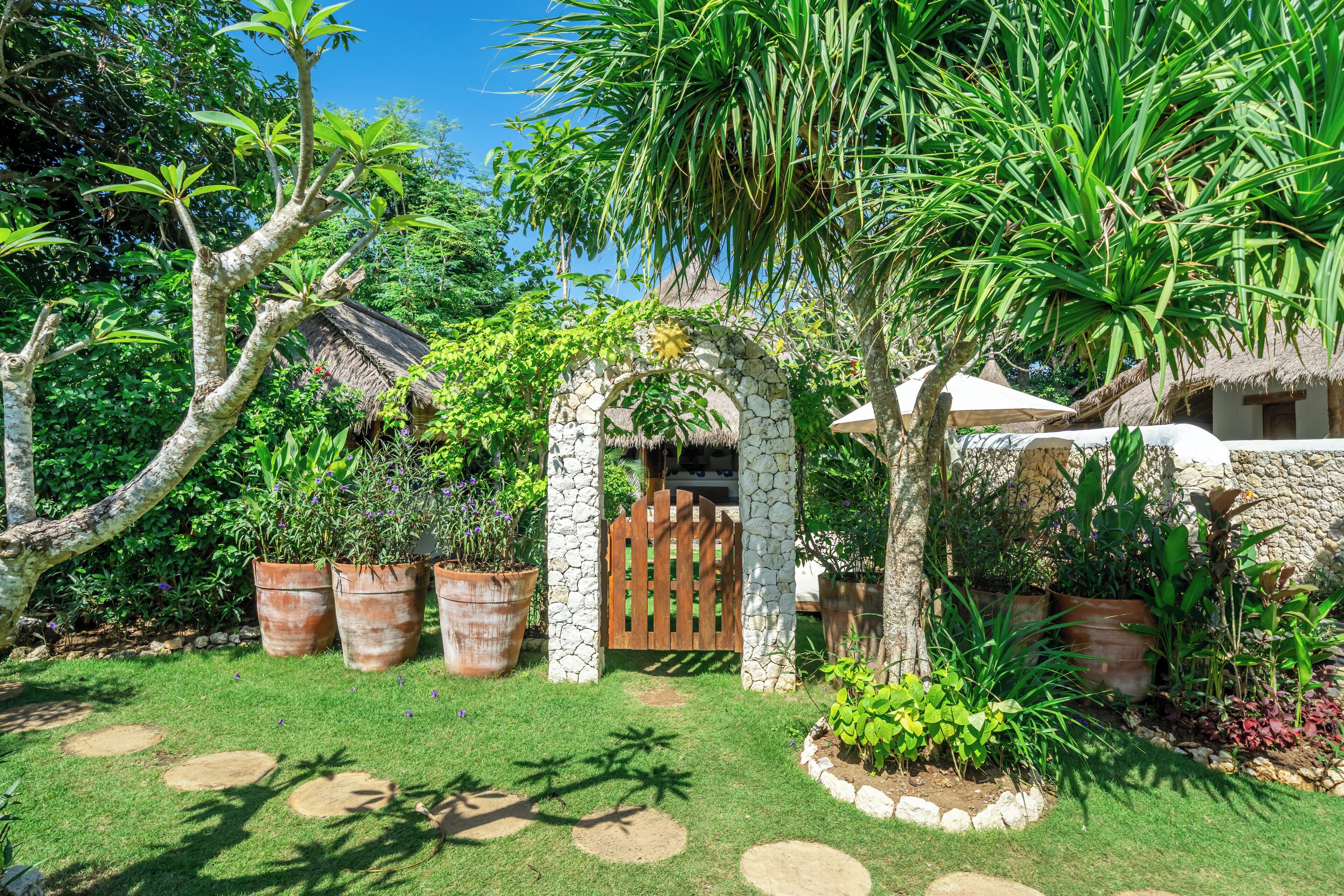 Um jardim tropical com um arco de pedra, plantas em vasos e uma construção com telhado de palha.