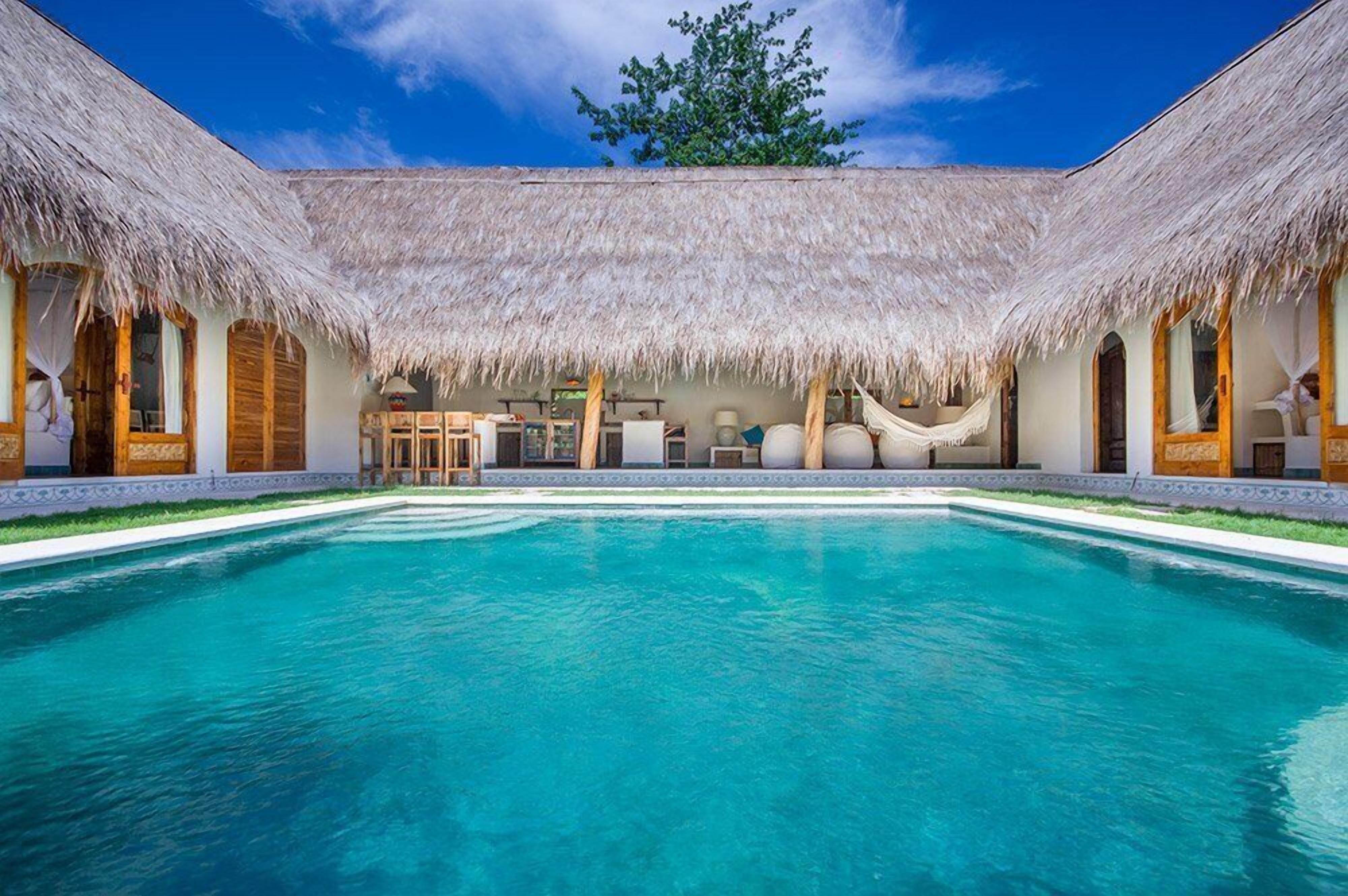 A poolside area with a thatched roof, a pool, and a hammock.