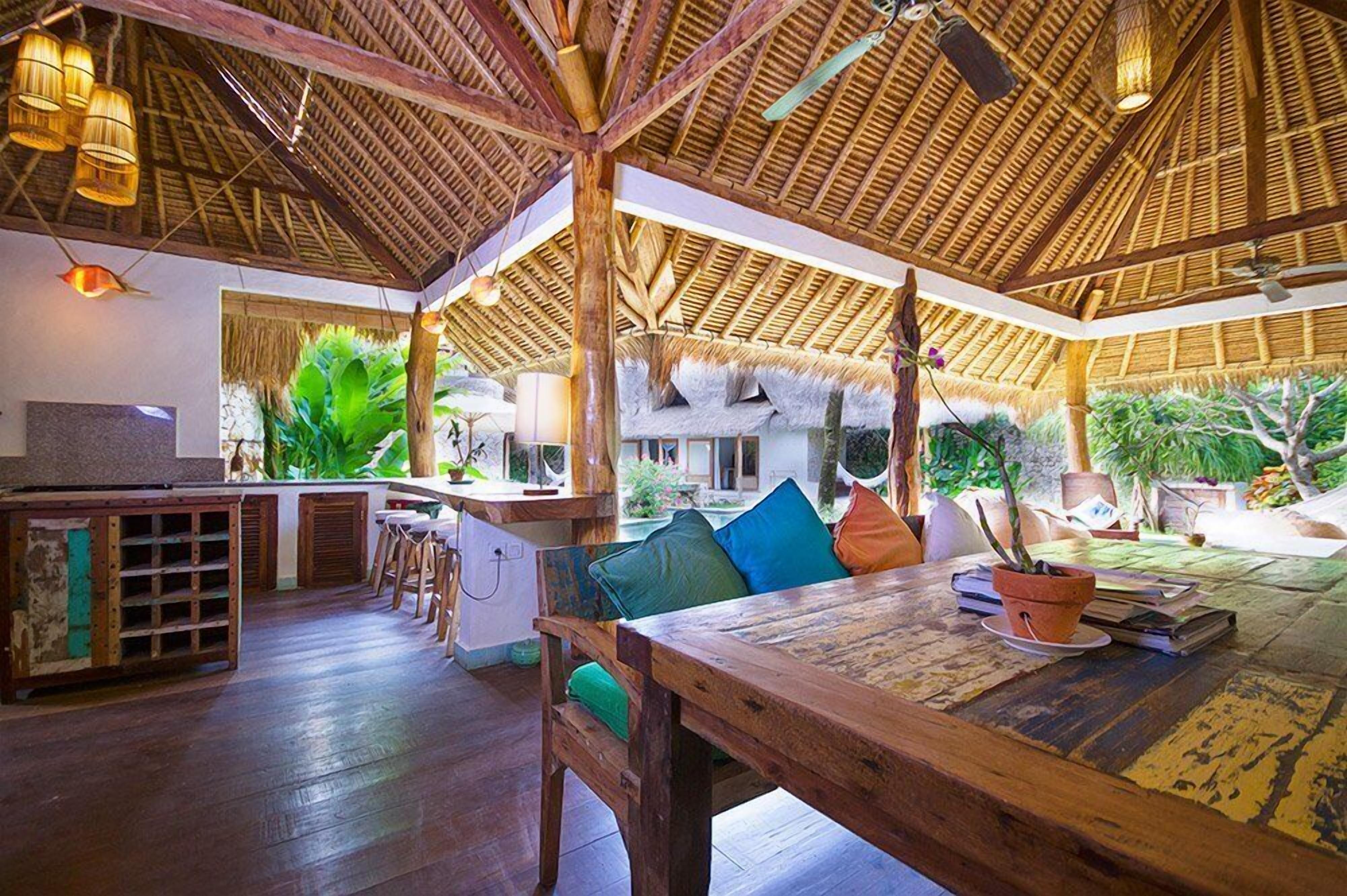 A spacious indoor area with a wooden ceiling and thatched roof elements, a dining table with chairs, and a bar area with stools.