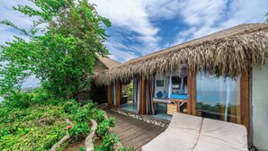 Un bungalow con tetto di paglia, un portico in legno, una veranda vetrata e un letto con materasso bianco.