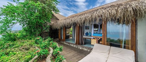 A thatched-roof bungalow with a wooden deck, a glass-enclosed room, and a bed with a white mattress.