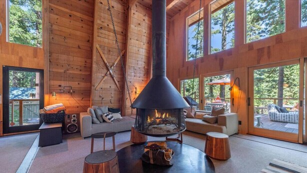 A cozy cabin interior with a central wood-burning stove, comfortable seating, and large windows offering views of the surrounding forest. - Classic Chamberlands Outdoor Living & Soaking Tub! (Homewood)