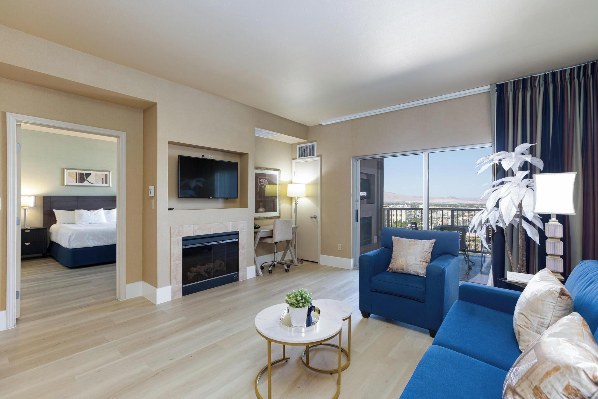 A modern hotel room with a living area, a bedroom, a fireplace, a TV, and a balcony with a view.