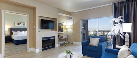 A modern hotel room with a living area, a bedroom, a fireplace, a TV, and a balcony with a view.