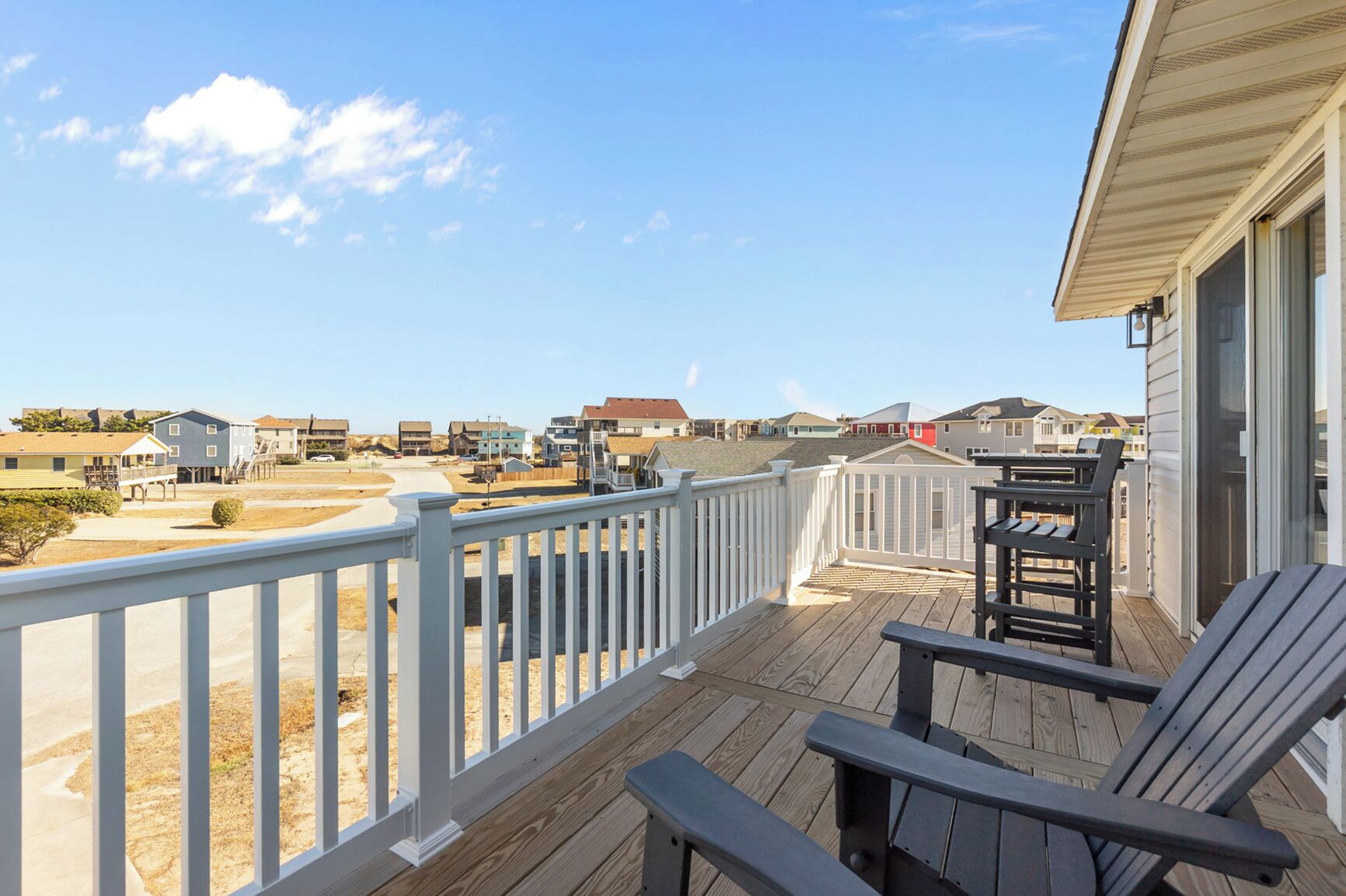 A wooden deck with a white railing, two lounge chairs, and a view of a residential area with houses and a clear sky.
