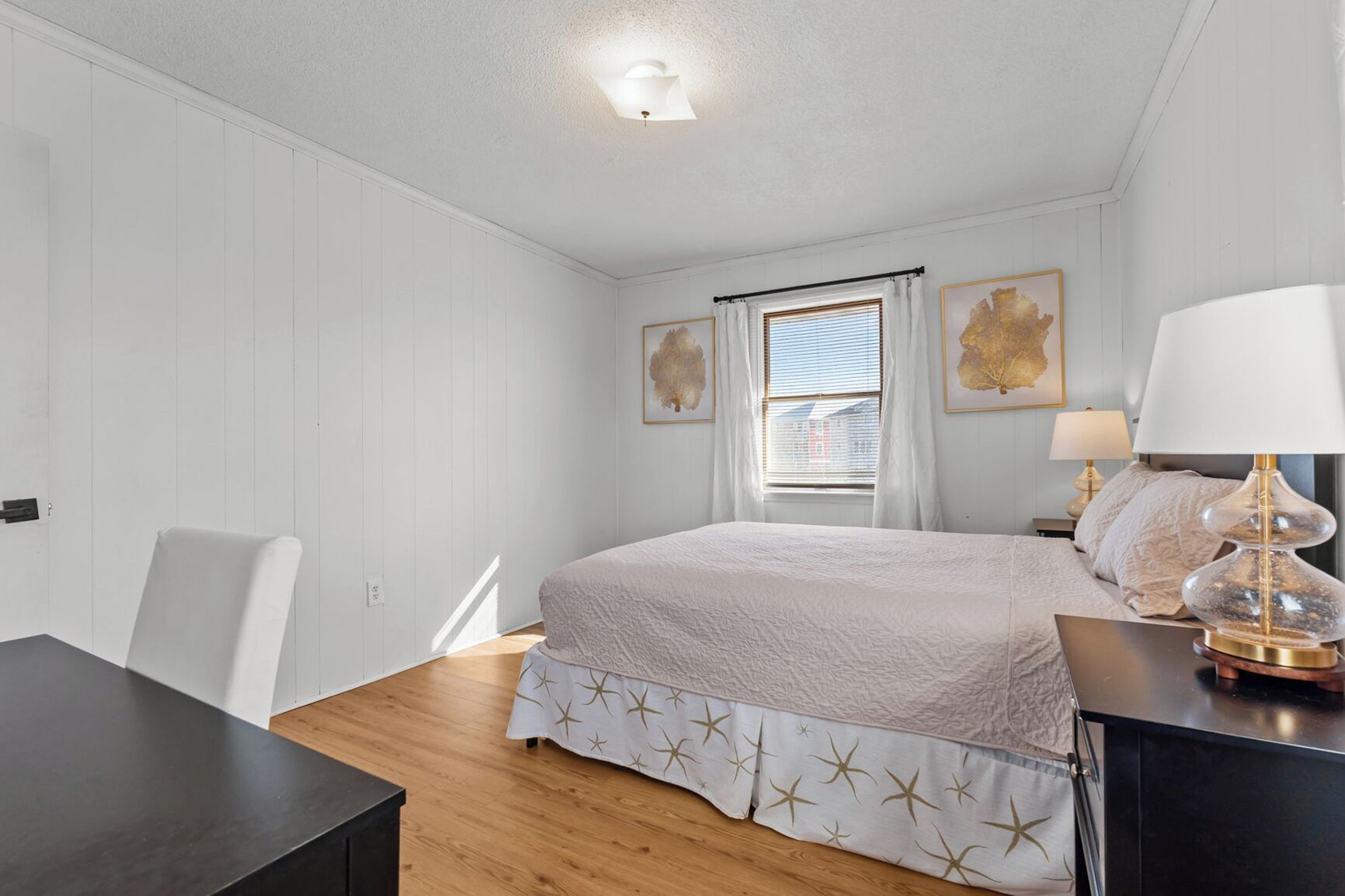 A bedroom with a bed, a desk, and a chair. There is a window with curtains and a lamp on a bedside table.