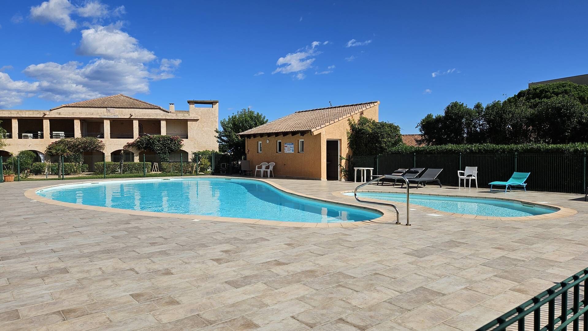 Une piscine à bord incurvé, entourée d’une terrasse dallée. On y trouve des chaises longues et un petit bâtiment avec un toit en tuiles, situé à proximité.