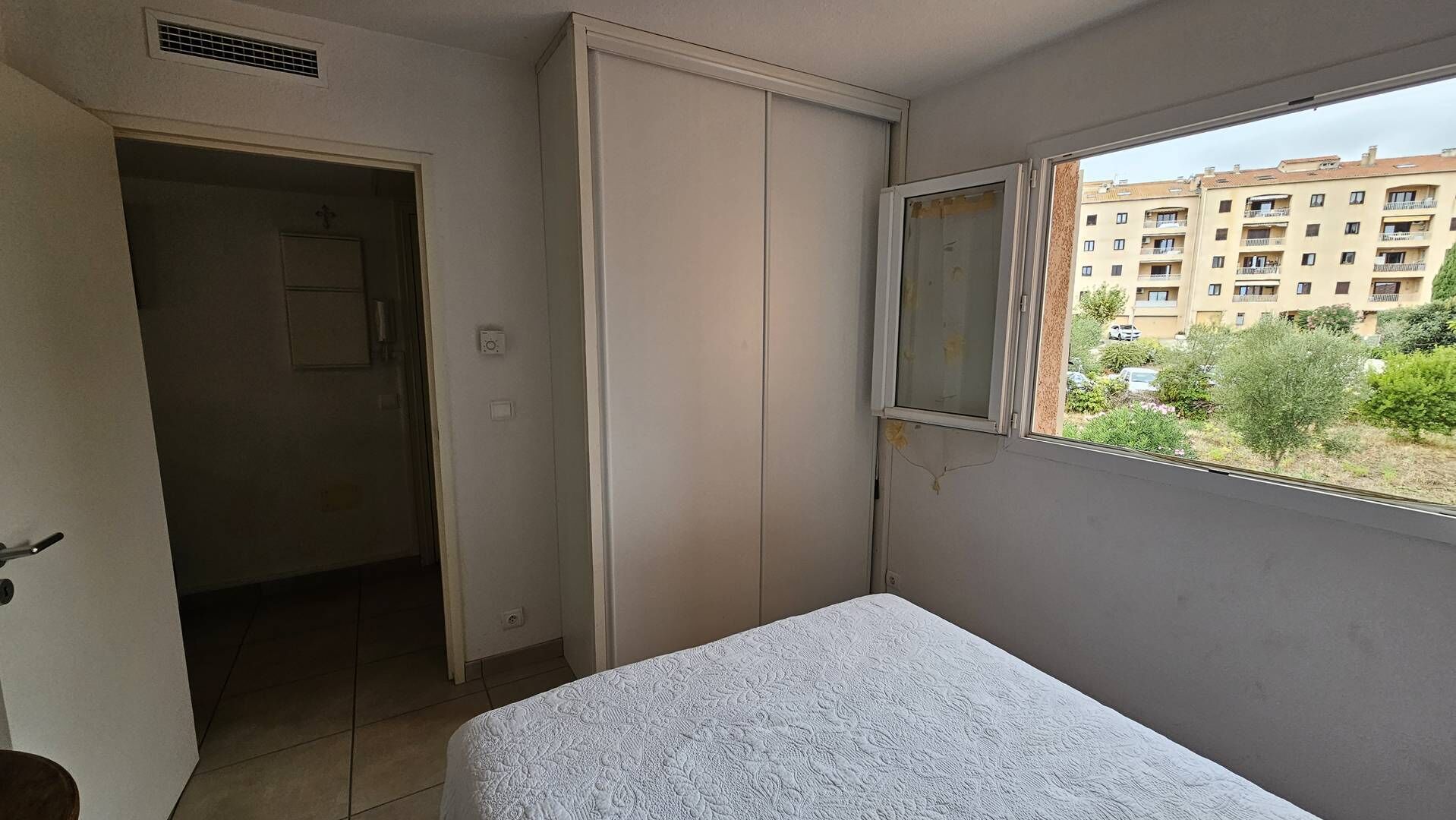 Een klein appartement met één kamer, een bed, een kledingkast en een raam met uitzicht op een woonwijk.