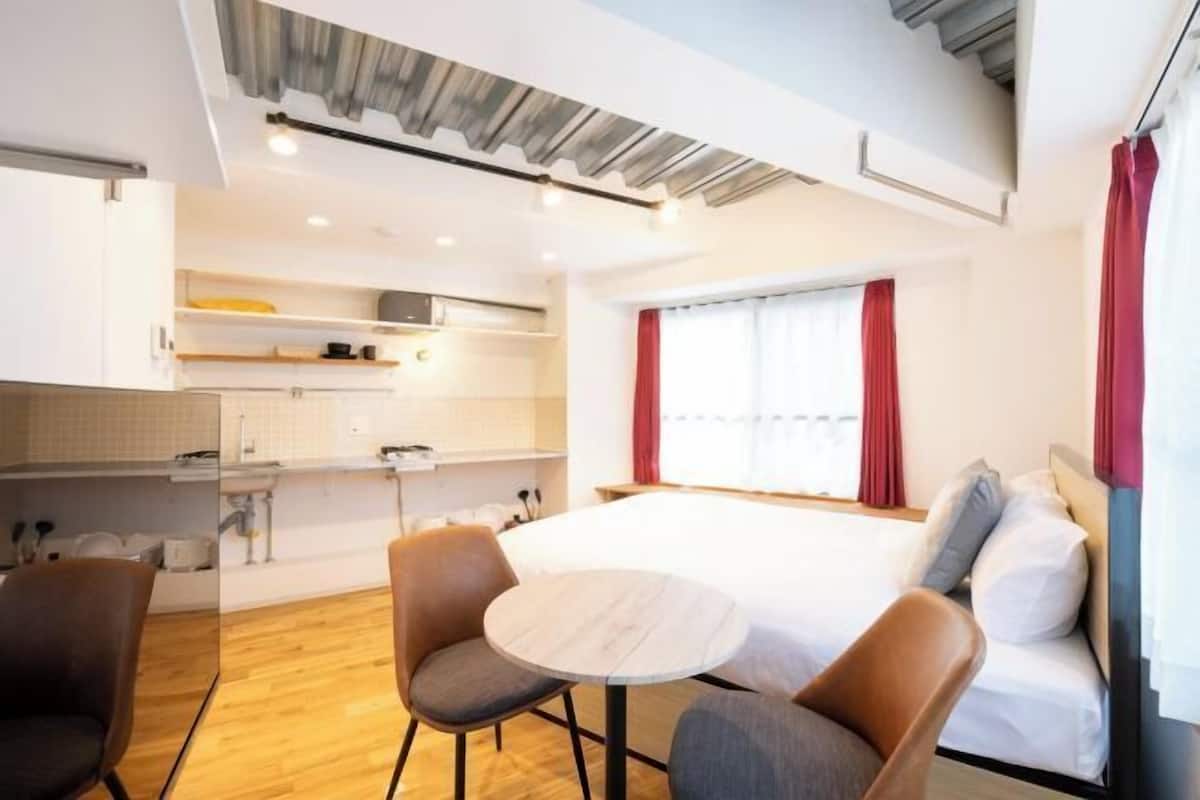 A modern hotel room with a bed, a small table, two chairs, a kitchenette, and a window with curtains.