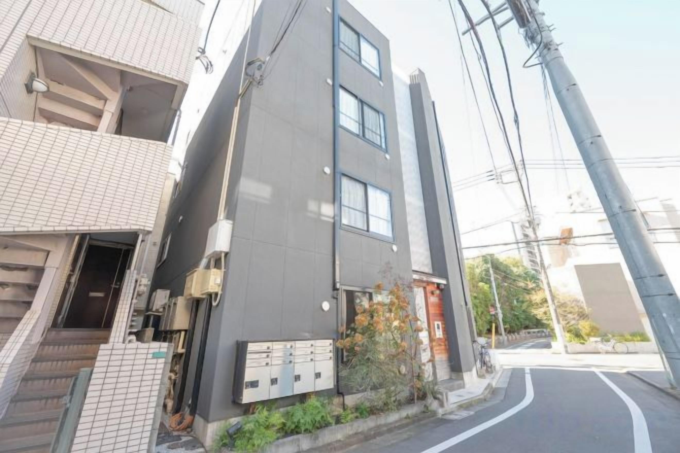 A multi-story building with a modern architectural design, featuring a staircase leading to an entrance, a small garden area, and a visible utility box.