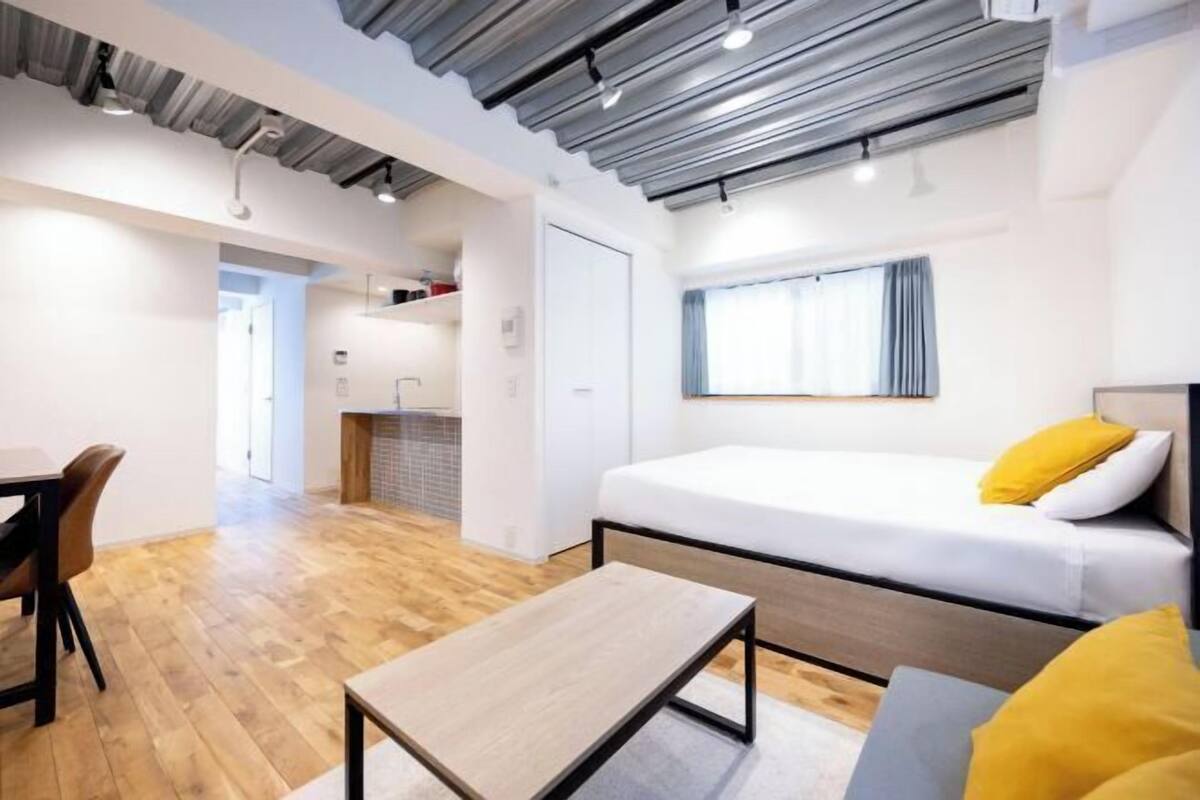 A modern room with a bed, a table, and a chair. There is a window with curtains, a door, and a kitchenette area.