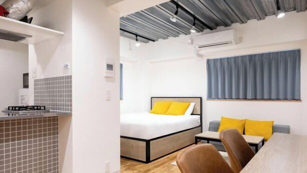 A compact hotel room with a kitchenette, a bed with yellow pillows, a dining area with a table and chairs, and a large window with a blue curtain. - Gran FusionIkebukuro (Tokyo)