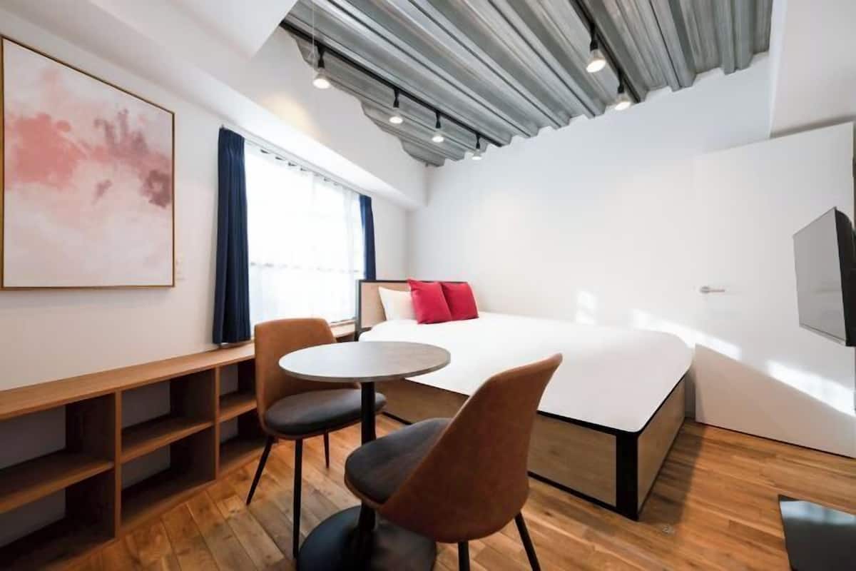 A modern hotel room with a bed, two chairs, a small table, a large abstract painting, and a window with curtains.