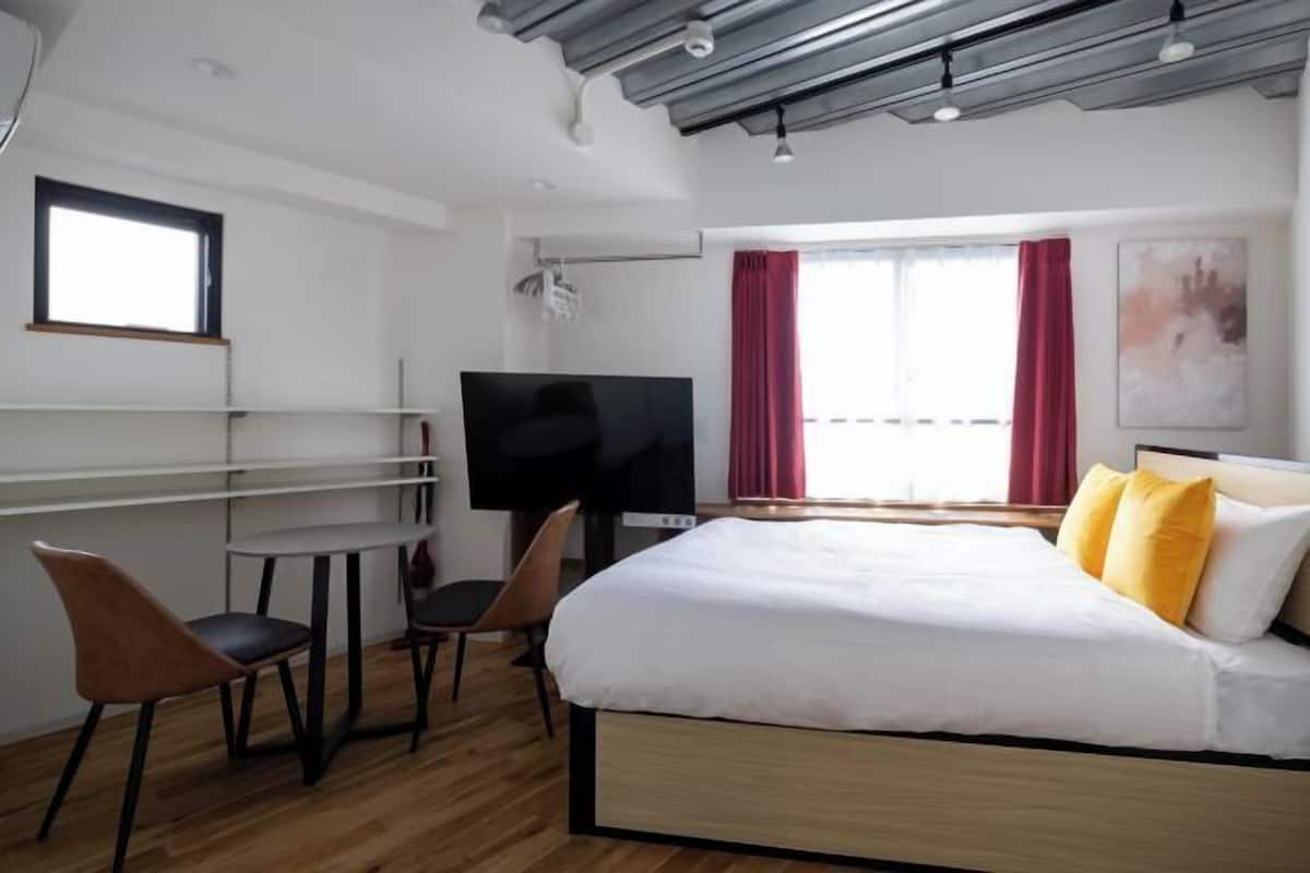 A modern hotel room with a bed, a desk with chairs, a TV, and a window with red curtains.
